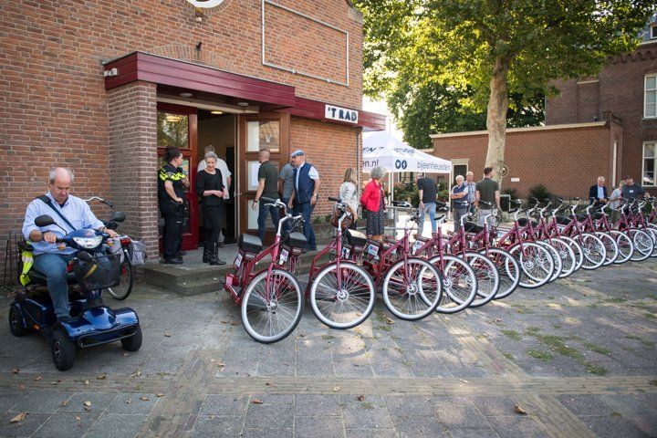 Mensen verzamelen zich bij een gebouw met een bordeauxrode luifel en fietsen. Een persoon op een scootmobiel rijdt ervoor.