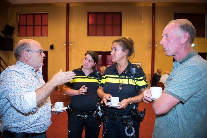 Twee politieagenten in gesprek met twee burgers binnenshuis, mogelijk in verband met een evenement.