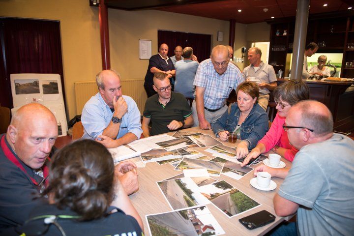 Een groep mensen bekijkt foto's op een ronde tafel, binnen. Sommigen praten en wijzen; anderen staan ​​er vlakbij.