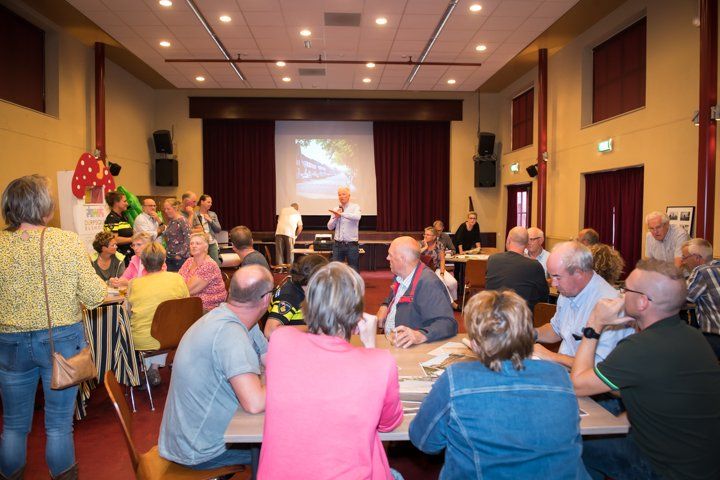 Mensen in een zaal: een presentatie op een scherm, een spreker, een zittend publiek aan tafels, rode gordijnen.