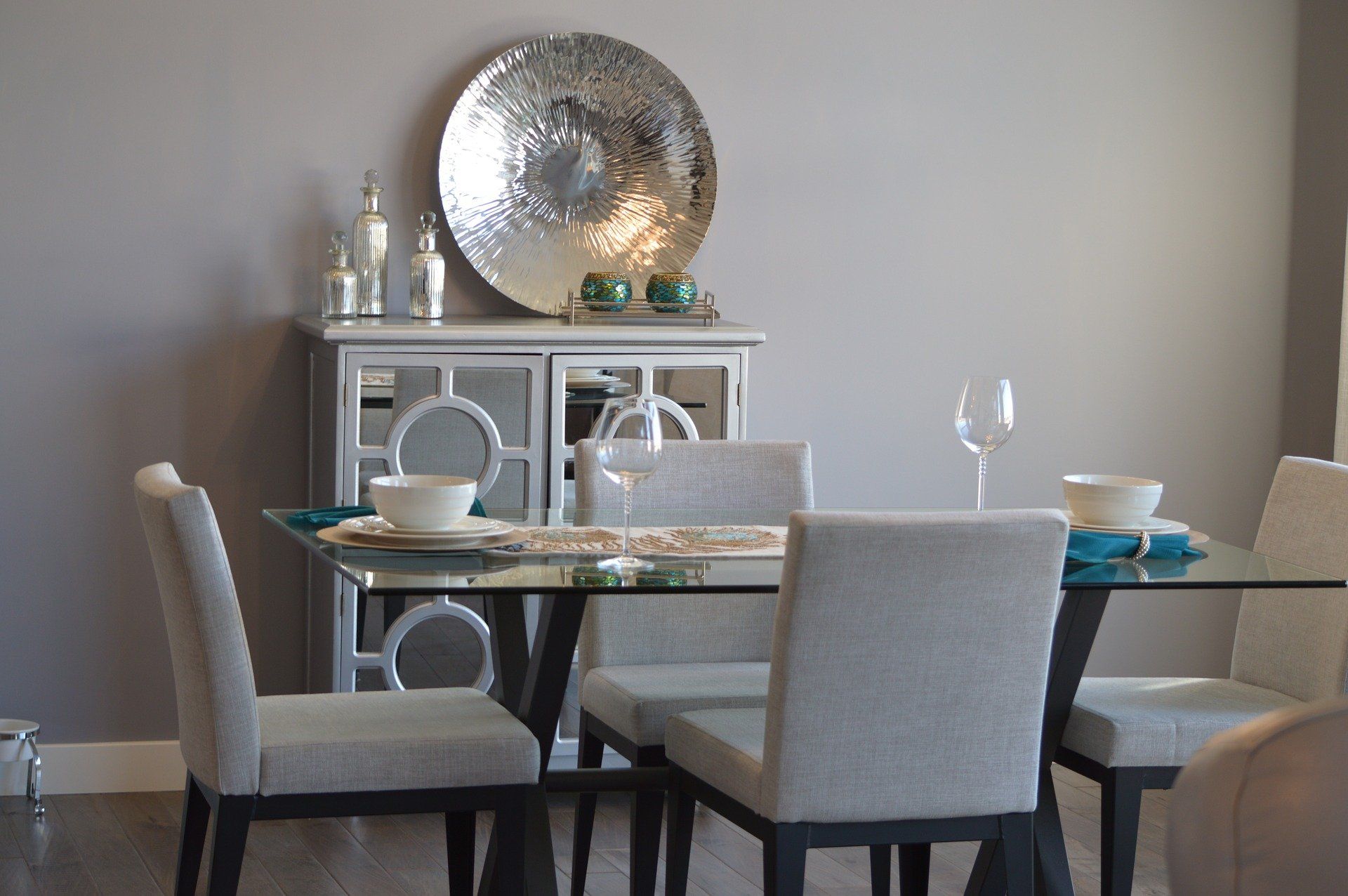 Dining room with glass table, gray chairs, mirrored cabinet, and decorative silver plate on the wall.