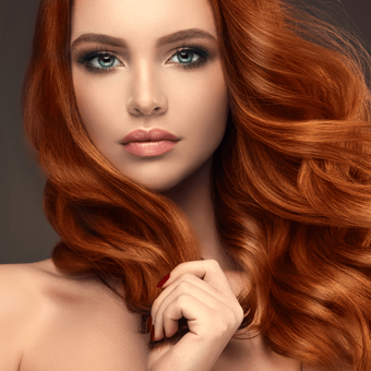 Woman with red wavy hair, smoky eye makeup, holding hair, fair skin, looking towards the viewer.
