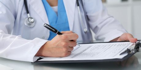 Doctor in white coat writing on clipboard with pen. Stethoscope around neck.