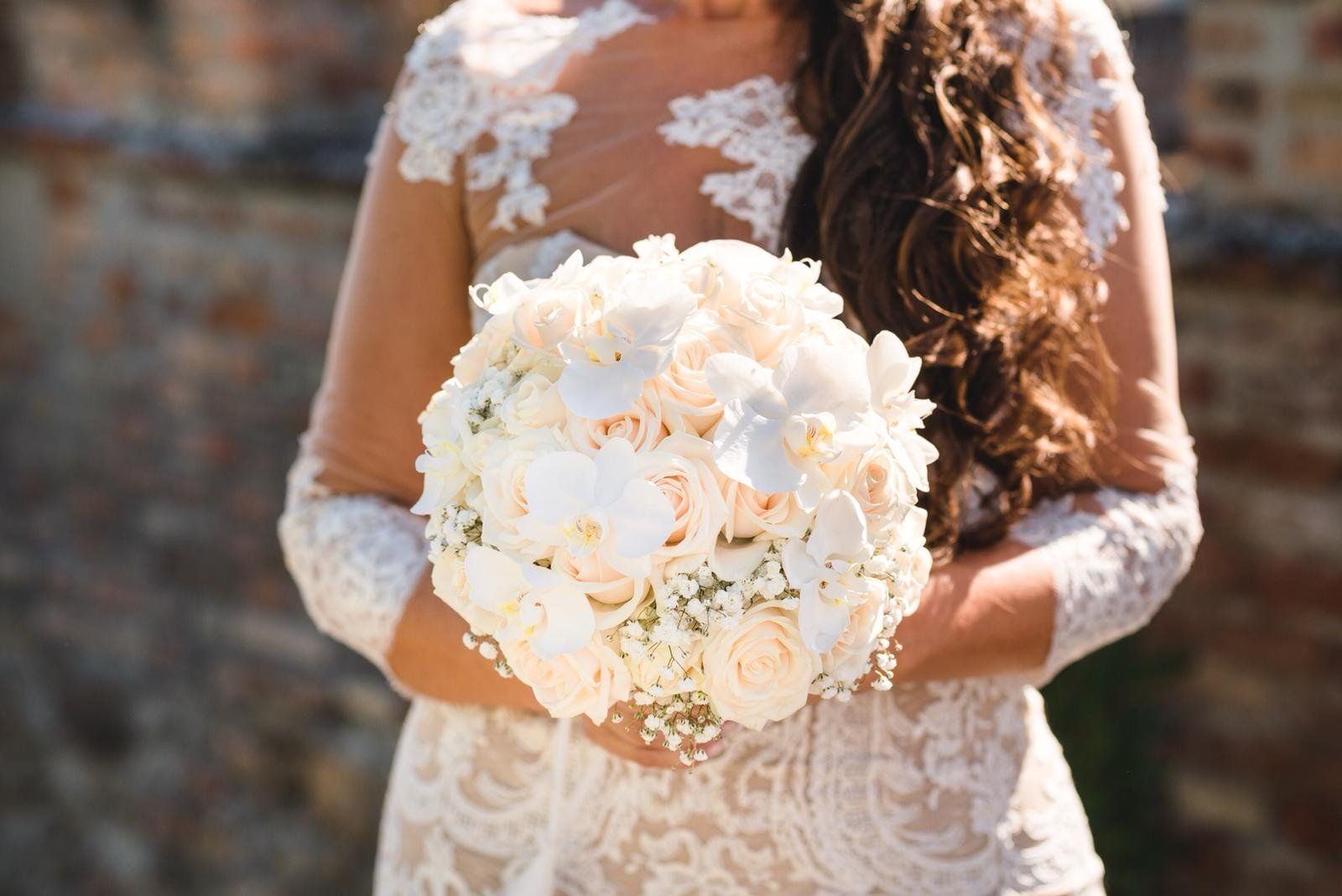 Sposa in abito di pizzo bianco che tiene in mano un bouquet di fiori bianchi all'aperto