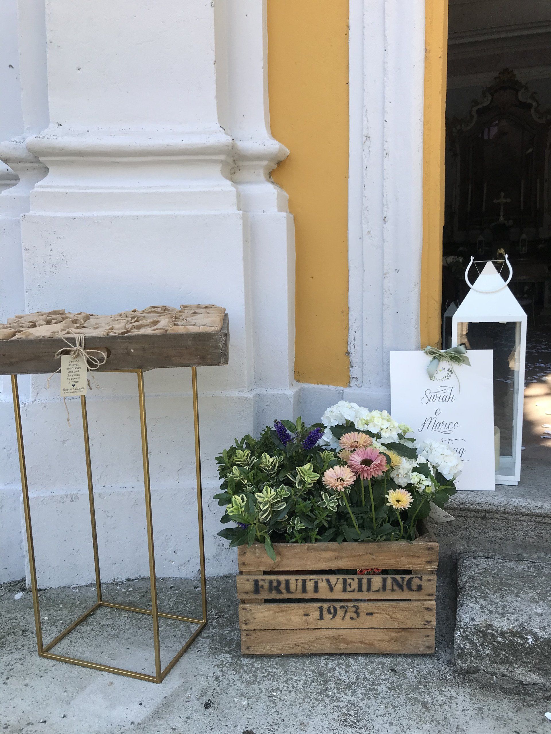 Composizione floreale in una cassa di legno accanto a un edificio bianco e a un supporto per insegna a forma di lanterna.