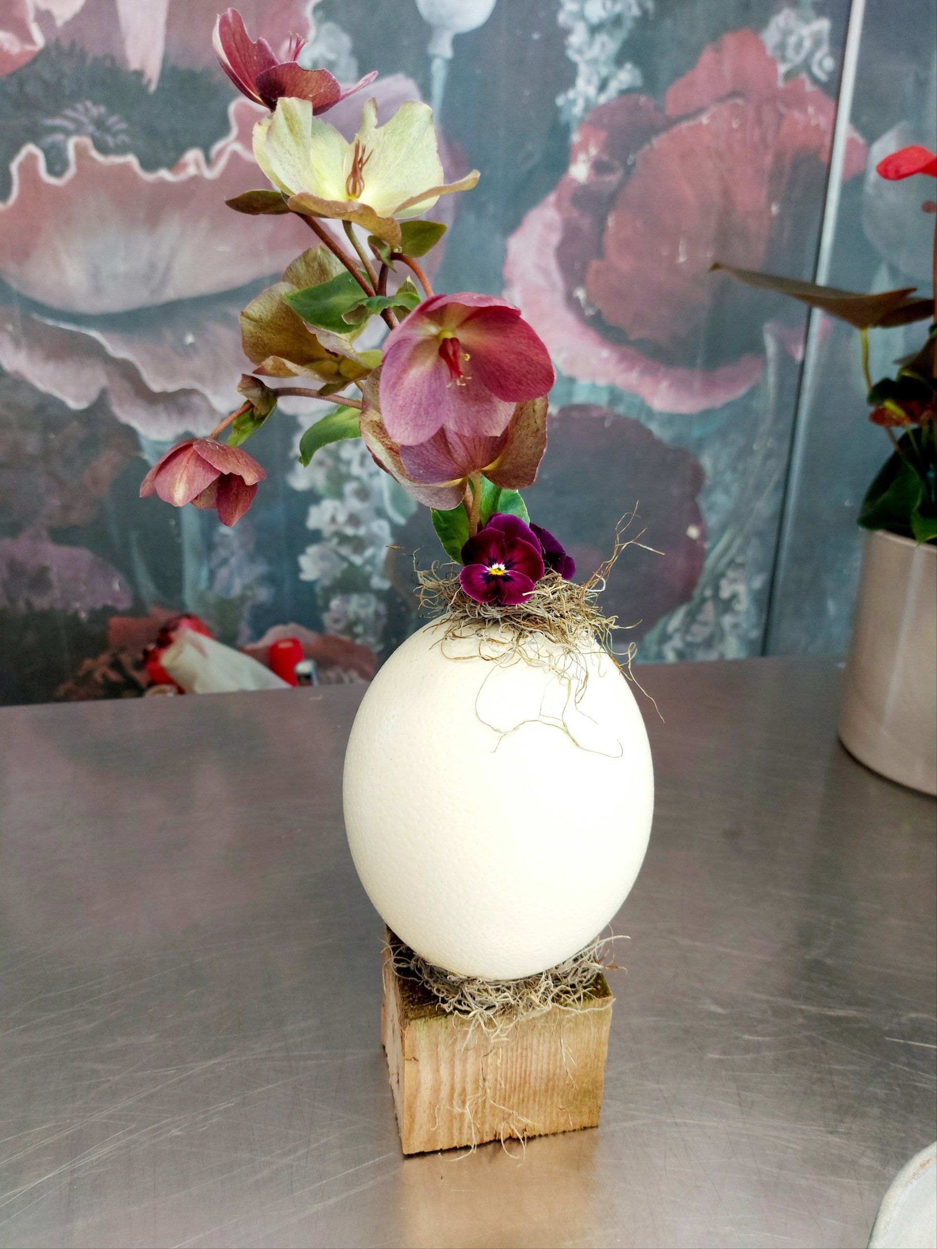 Vaso bianco a forma di uovo con fiori viola su un blocco di legno, su uno sfondo floreale.