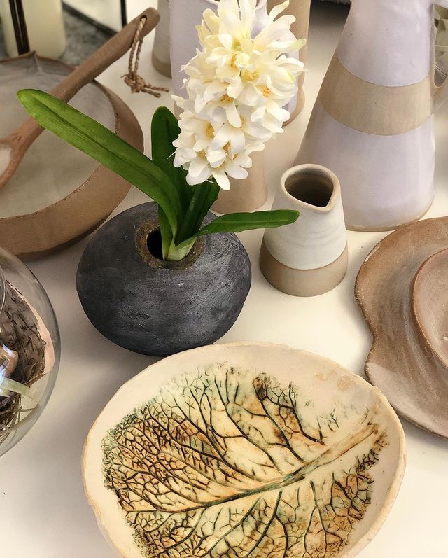 Fiore bianco in vaso nero con decorazioni in ceramica e piatto con motivo a foglie su un piano del tavolo beige.