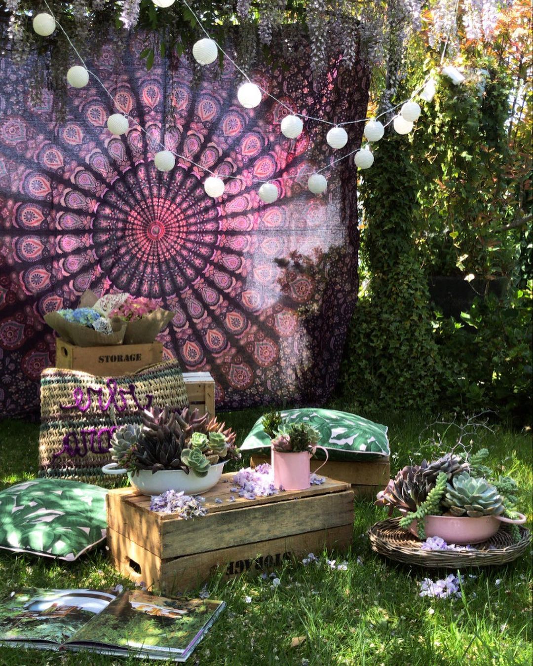 Arredamento da giardino in stile boho con sfondo di arazzo rosa, luci decorative e tavolini bassi in legno sull'erba.