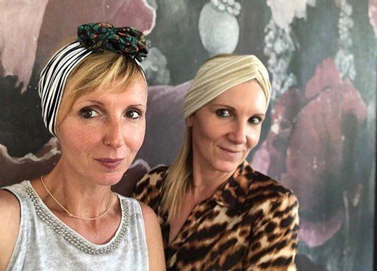 Due donne in posa in interni, sorridenti verso la macchina fotografica davanti a uno sfondo floreale.
