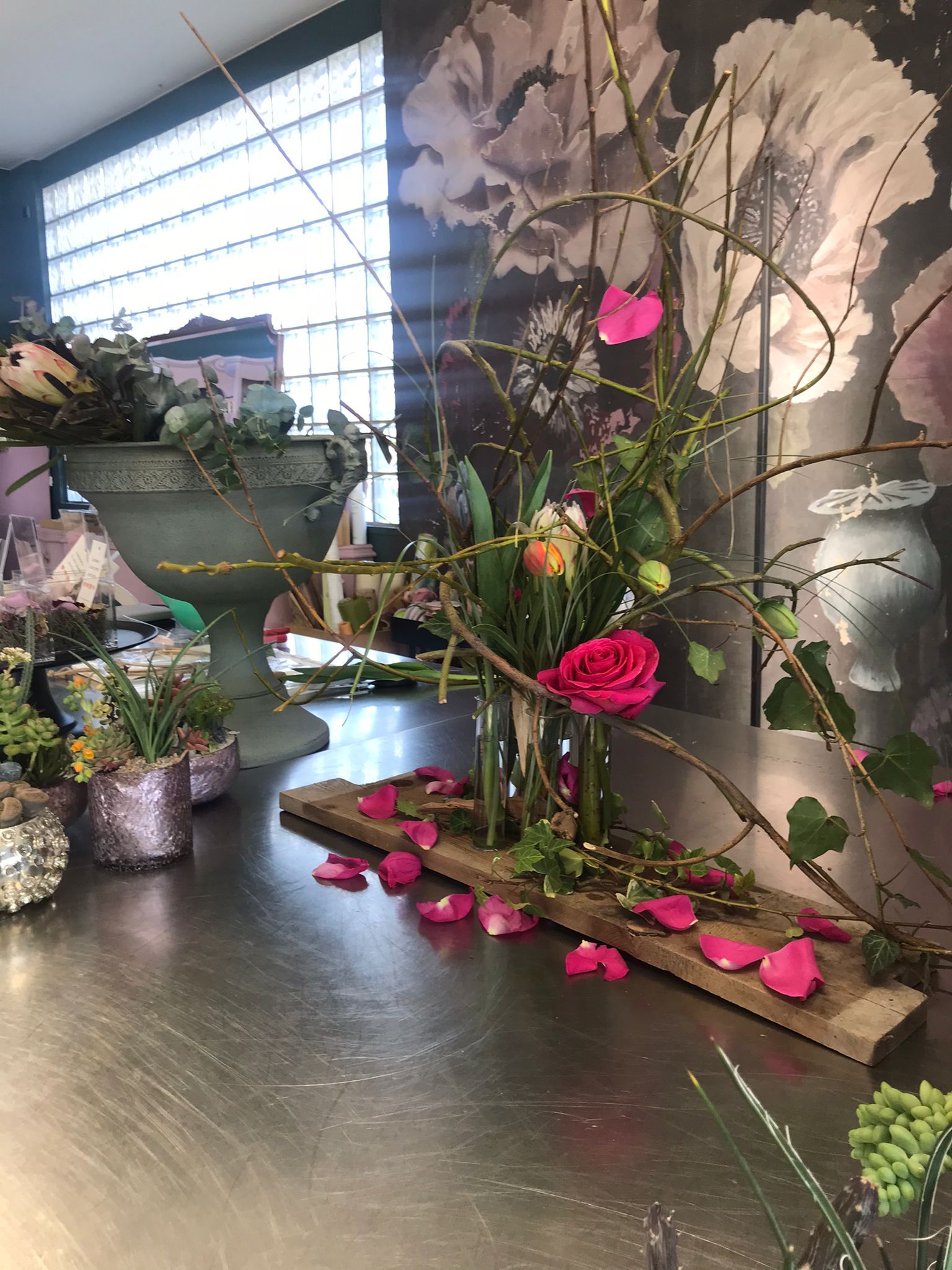 Composizione floreale con fiori e rami rosa su un tavolo accanto a una finestra luminosa e a un murale.
