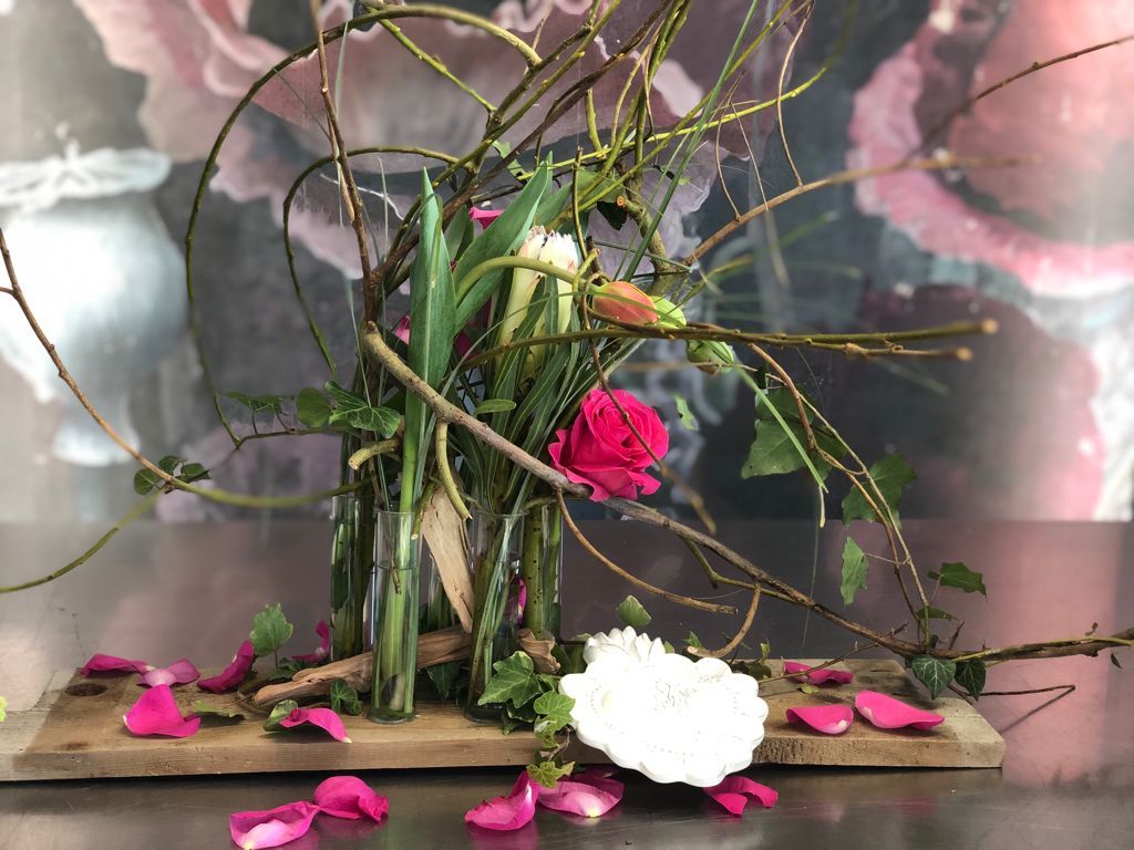 Composizione floreale con fiori rosa e bianchi, rami intrecciati e petali sparsi su una base di legno.