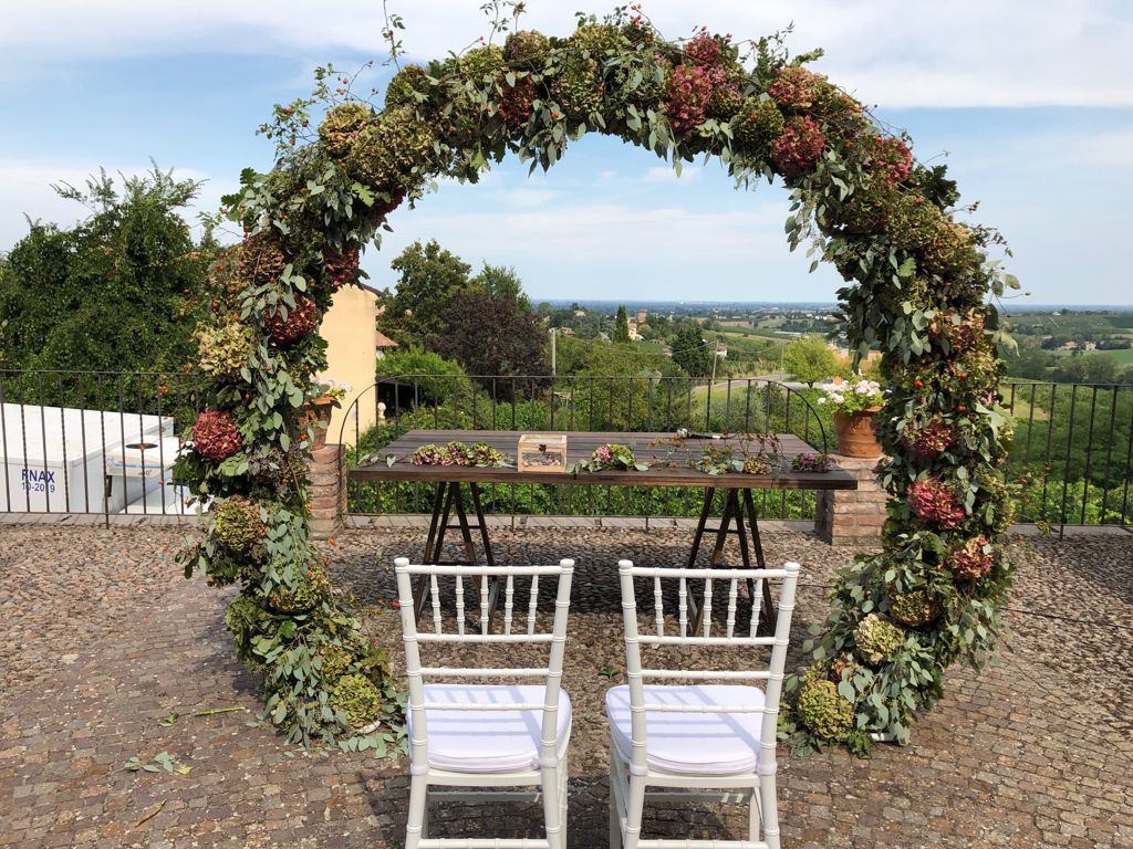 Sedie bianche davanti a un arco ricoperto di fiori che si affaccia su un pittoresco paesaggio vitivinicolo.