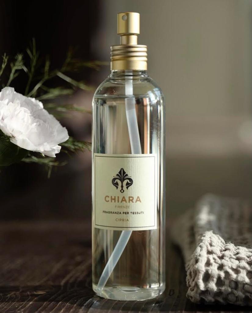 Flacone di profumo trasparente con vaporizzatore dorato su una superficie scura, accanto a fiori bianchi e decorazioni.