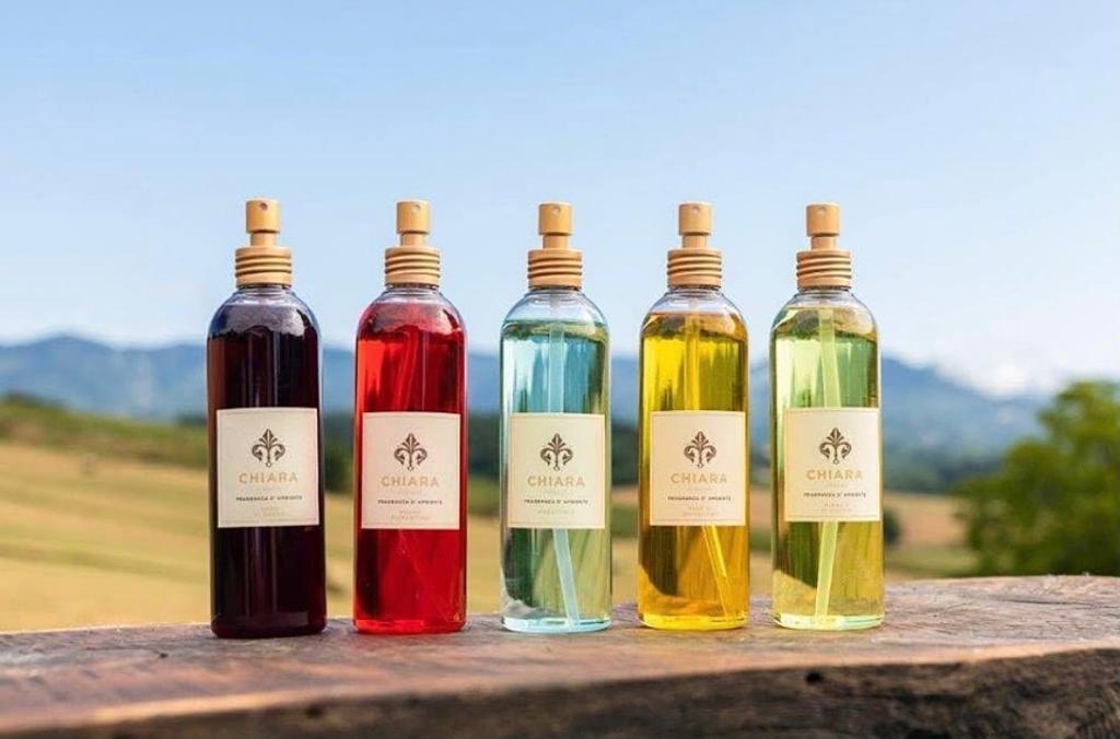 Cinque bevande colorate in bottiglia su una mensola di legno, con uno sfondo sfocato di paesaggio esterno.