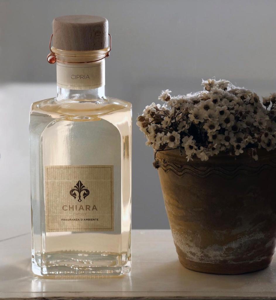 Profumo trasparente accanto a un piccolo vaso di fiori marrone con fiori bianchi secchi su una superficie chiara.