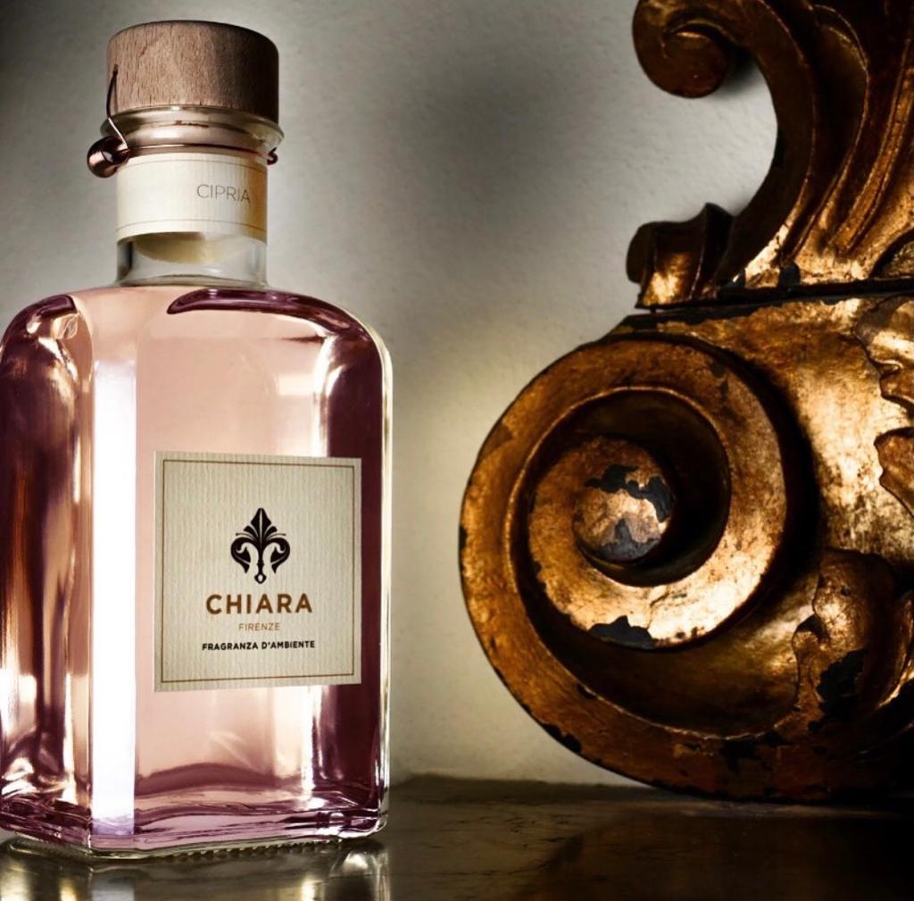 Flacone di profumo rosa con l'etichetta CHIARA accanto a un ornamento dorato intagliato su una superficie riflettente