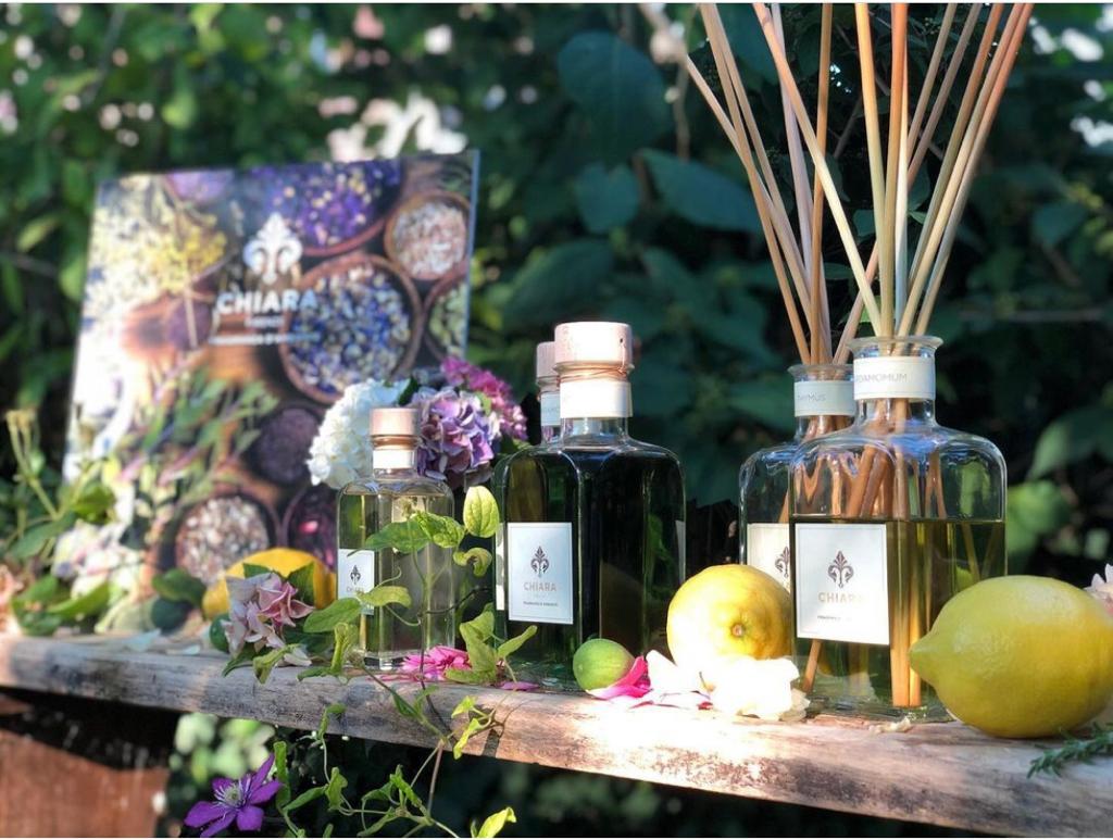 Esposizione di profumi botanici con flaconi di vetro, agrumi e fiori su un tavolo illuminato dal sole.