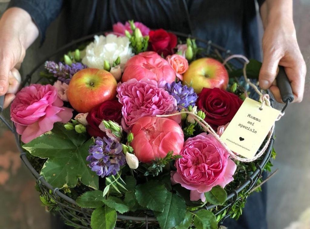 Cesto regalo a forma di cuore con frutta e fiori, rose rosa e mele, tenuto da due mani.