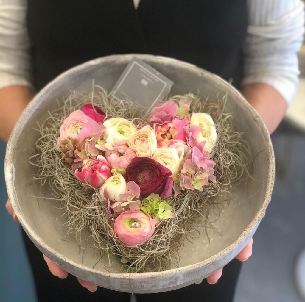 Una ciotola di fiori rosa e crema, composta da muschio, viene tenuta in mano da una persona con una maglietta nera.