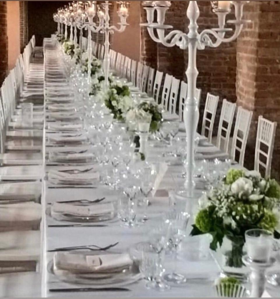 Un tavolo da banchetto apparecchiato con sedie bianche, centrotavola floreali e candelabri d'argento in una sala in mattoni.