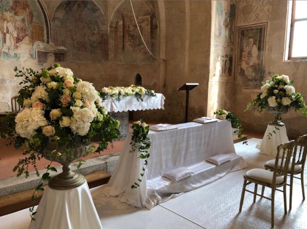 Altare nuziale con composizioni floreali bianche, tavolo drappeggiato e sedie in una rustica cappella in pietra.