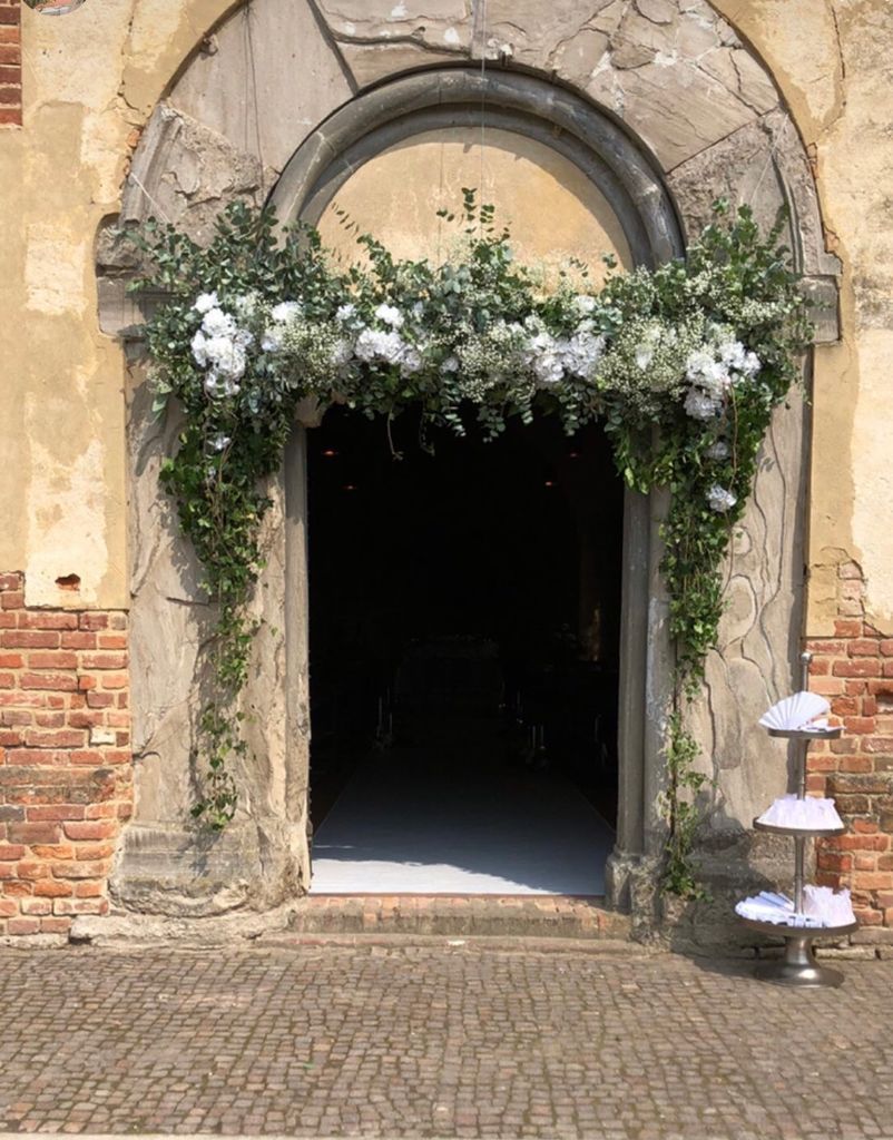 Antico portale in pietra con ingresso ad arco e fiori bianchi, che si apre su un interno buio.