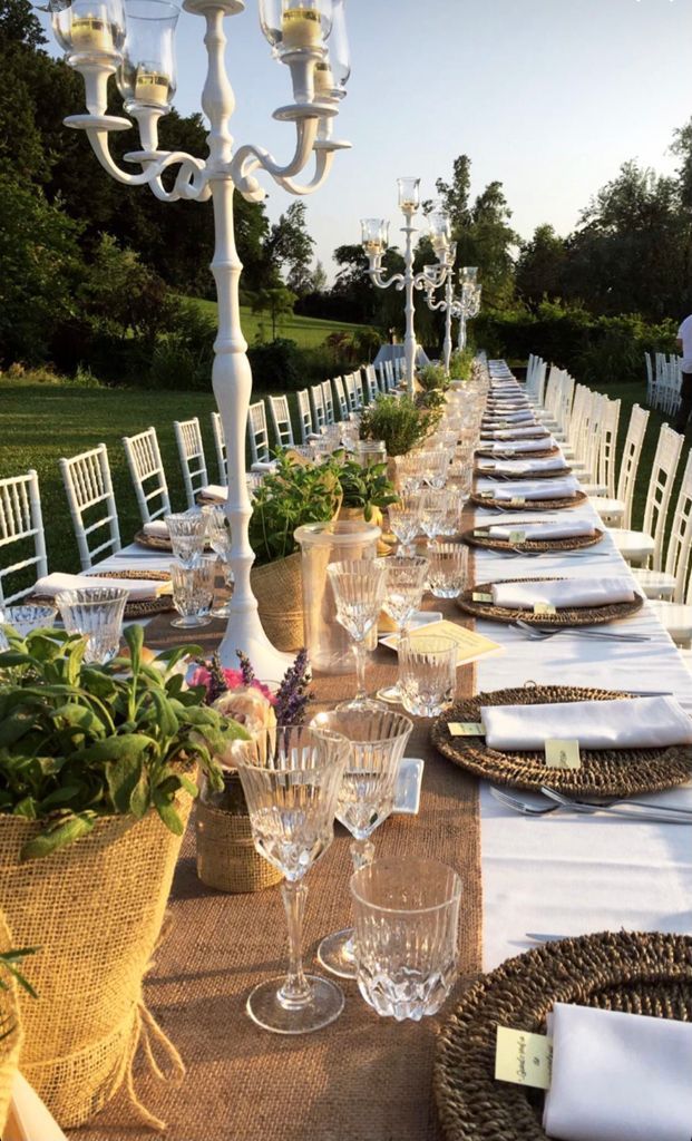 Un lungo tavolo da pranzo all'aperto con sedie bianche, candele e fiori in un giardino al tramonto.