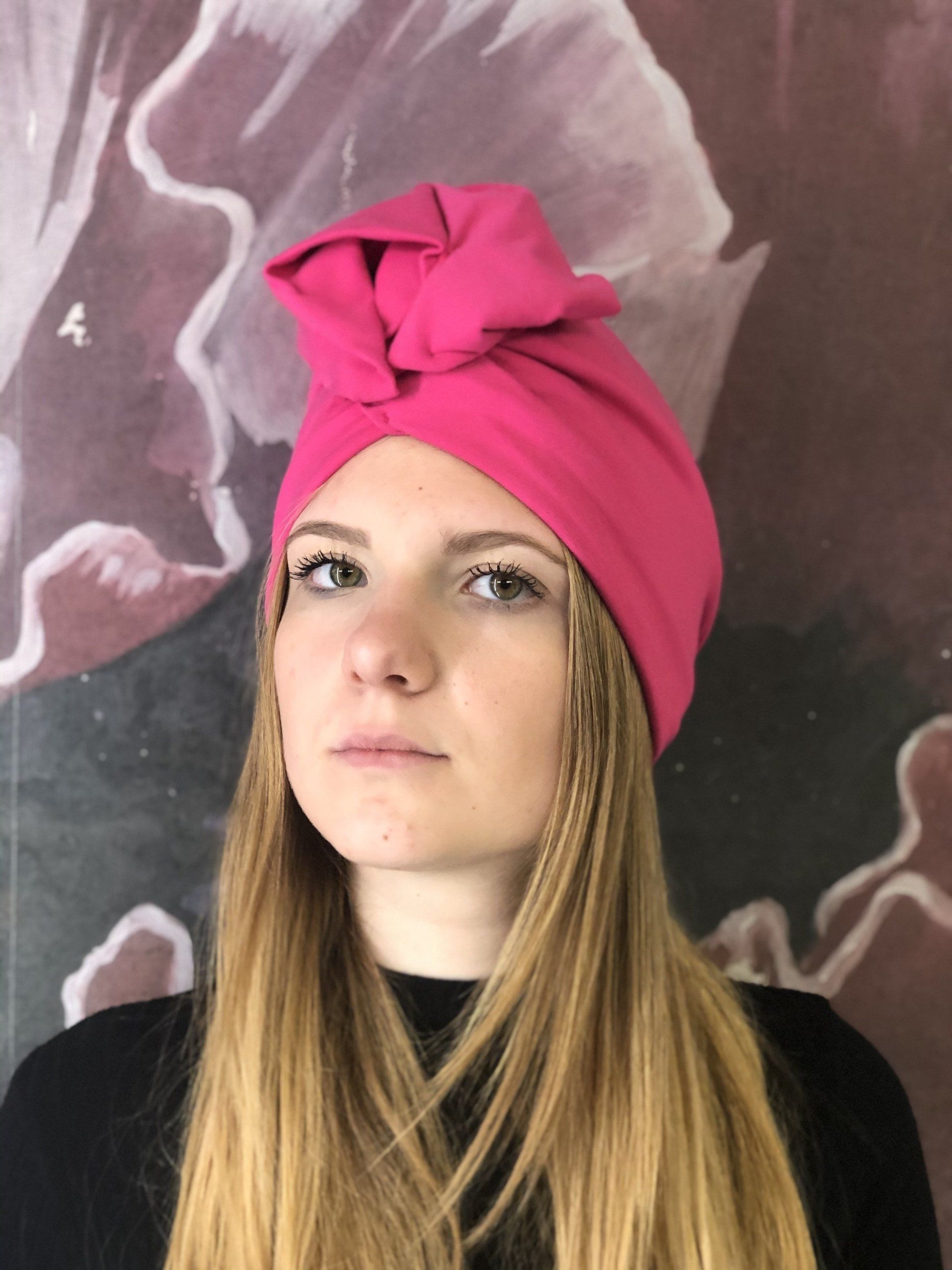 Ritratto di una persona con un turbante rosa acceso su uno sfondo di un murale astratto.