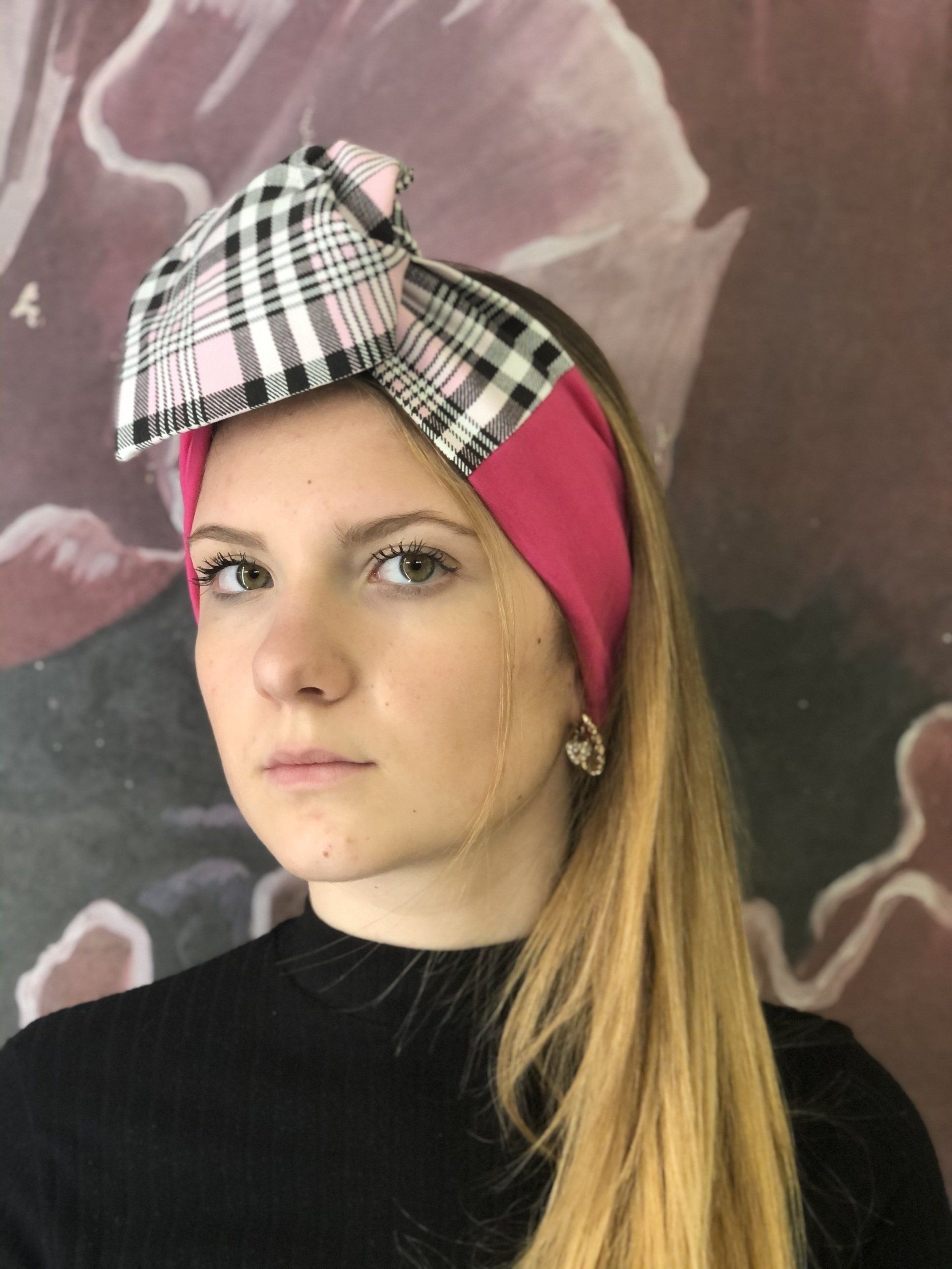 Ritratto di una persona con un foulard rosa e un fiocco a quadri, dall'espressione neutra, su sfondo scuro.