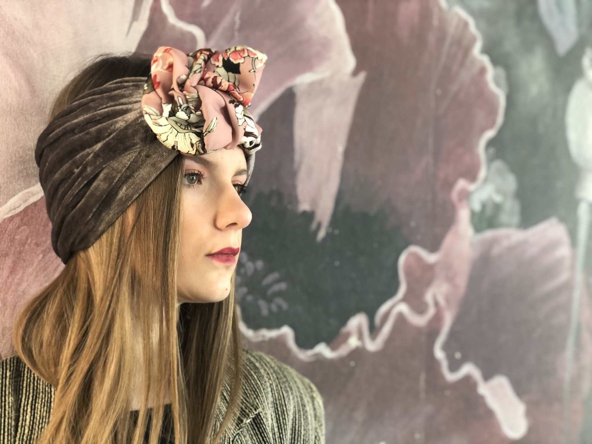 Donna con turbante di lana e fiocco floreale, rivolta a sinistra, sullo sfondo di un murale rosa tenue.