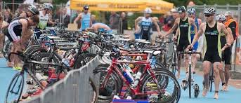 Triatleten in een wisselzone met hun fietsen. Atleten stappen op en af. De zone heeft fietsen en een blauwe vloer.