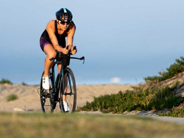 Triatleet met helm en zonnebril rijdt op een zwarte fiets op een pad langs zandduinen en groen.