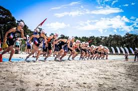 Een groep triatleten rent over een zandstrand richting de startlijn. Stralend blauwe lucht.