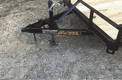 Black utility trailer hitch on gravel beside a pickup truck, with a metal ramp and jack stand.