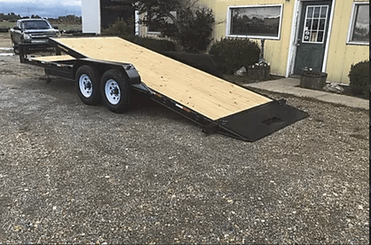 Flatbed trailer with plywood ramp parked on gravel beside a yellow building.