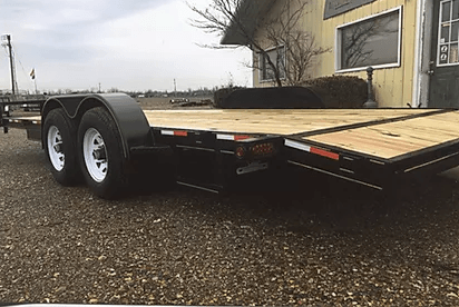 Black flatbed utility trailer with wooden deck and dual axles parked on gravel beside a building