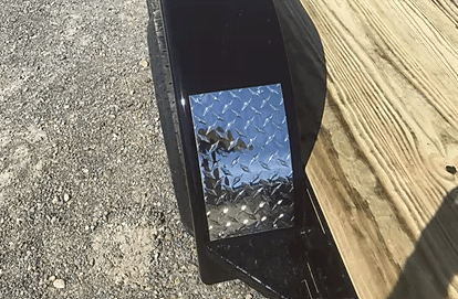 Close-up of a black vehicle side mirror reflecting pavement and a wooden fence in sunlight