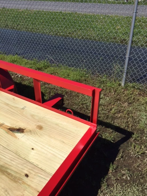 Red trailer with a wooden deck beside a chain-link fence and grassy roadside.
