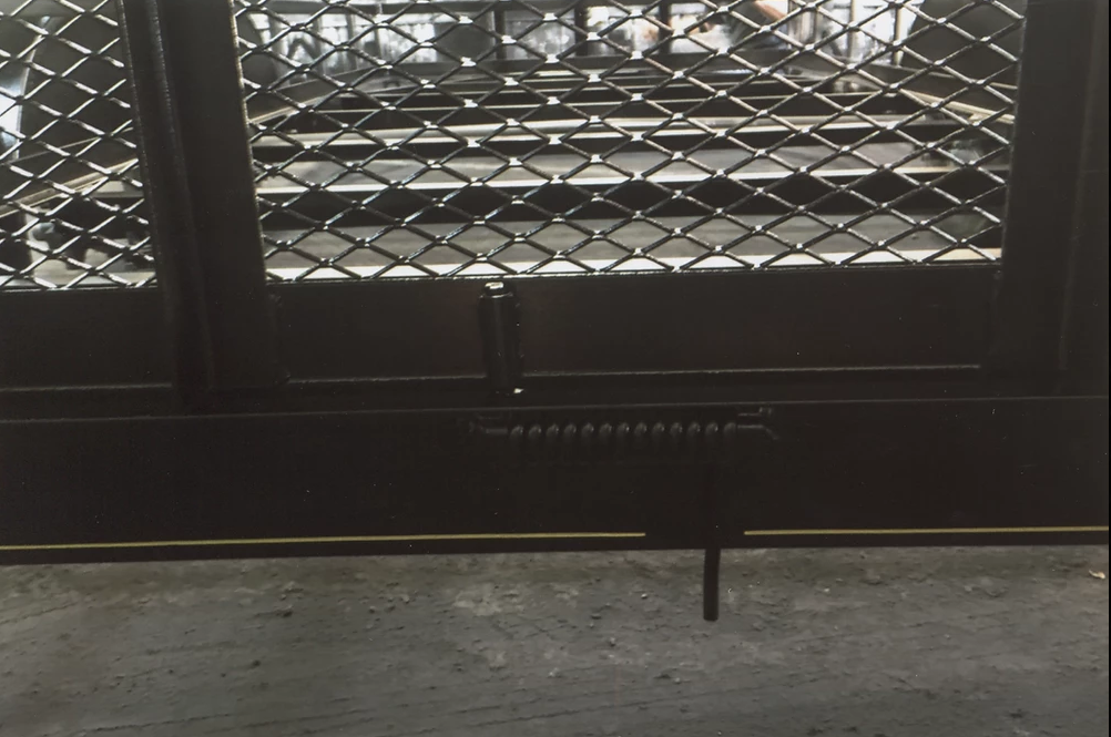 Dark metal gate with mesh panels, viewed from behind, over a concrete floor.