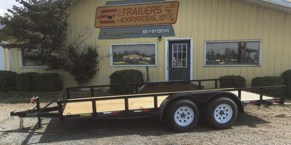 Flatbed utility trailer parked outside a trailer repair shop with a tan building and sign.