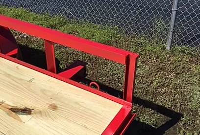 Red utility trailer with a wooden plank deck parked beside a chain-link fence.