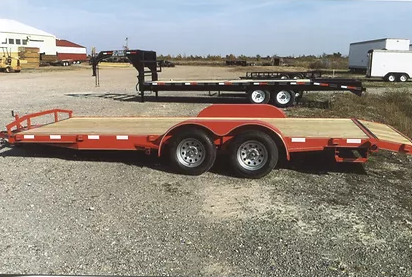 Red and black flatbed trailer parked on gravel in an open lot under a clear sky.