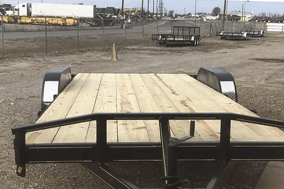 Black flatbed trailer parked on a dirt lot with industrial buildings in the background