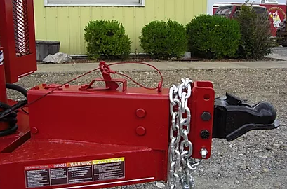 Red trailer hitch with safety chains attached, beside a yellow building and shrubs.