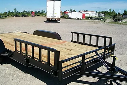 Black utility trailer with wooden deck parked in a gravel lot near trucks and trees