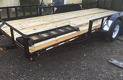 Black utility trailer with wooden deck and ramps parked on wet pavement.