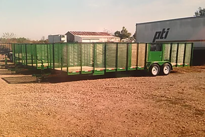Green mesh utility trailer with two wheels parked on a gravel lot beside a PTI building.