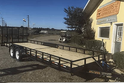 Black utility trailer parked beside a yellow building on a gravel lot under a clear sky
