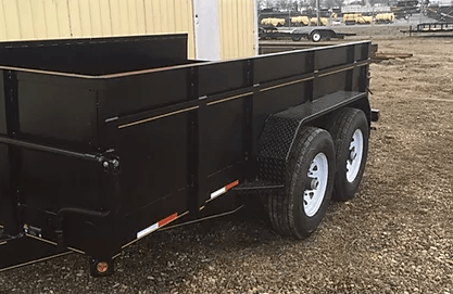 Black utility trailer with white wheels parked beside a building on gravel.