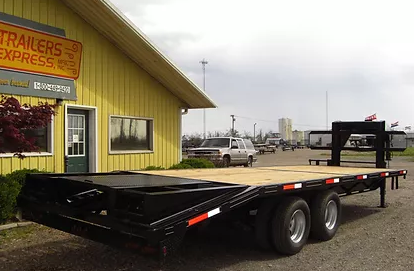 Black flatbed trailer parked beside a yellow trailer shop with industrial buildings in the background
